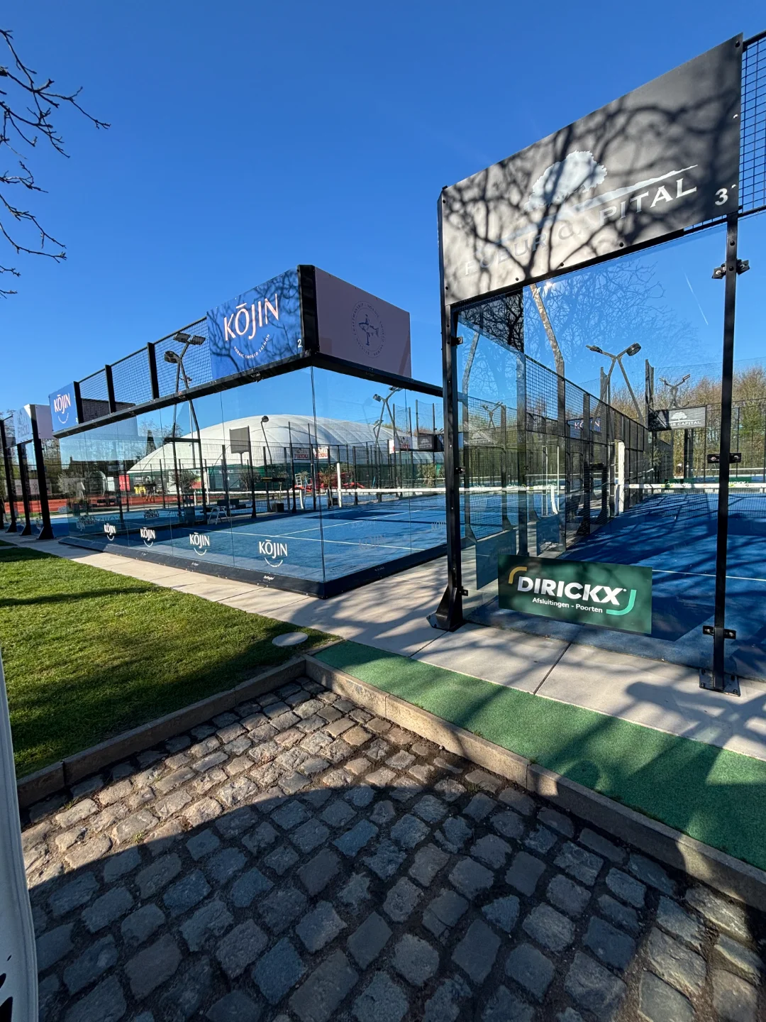 Padelbaan glaswanden & oprit reinigen – Renovivo diensten Schoon ogende padelbanen met glazen wanden en kasseien oprit in de zon, ideaal voor reiniging van terrassen en ramen door Renovivo.